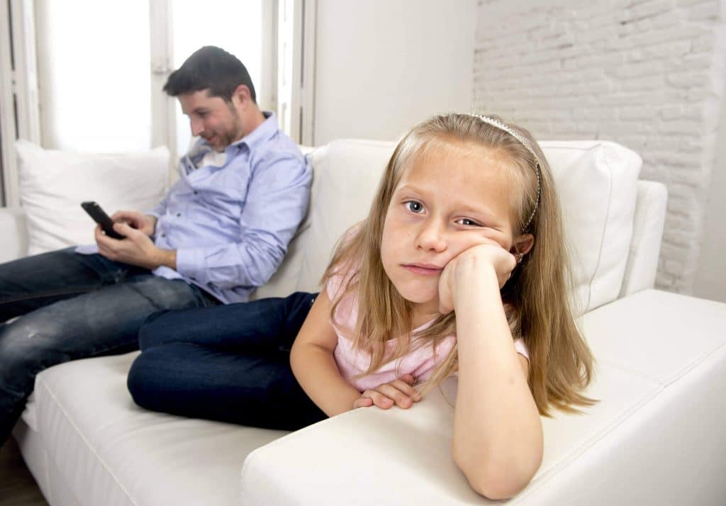 parent busy on smartphone while the little girl sits on the couch bored