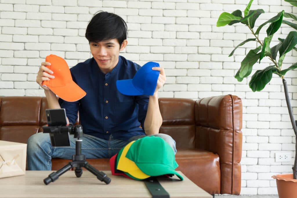 man holding two hats in front of a phone on a tripod