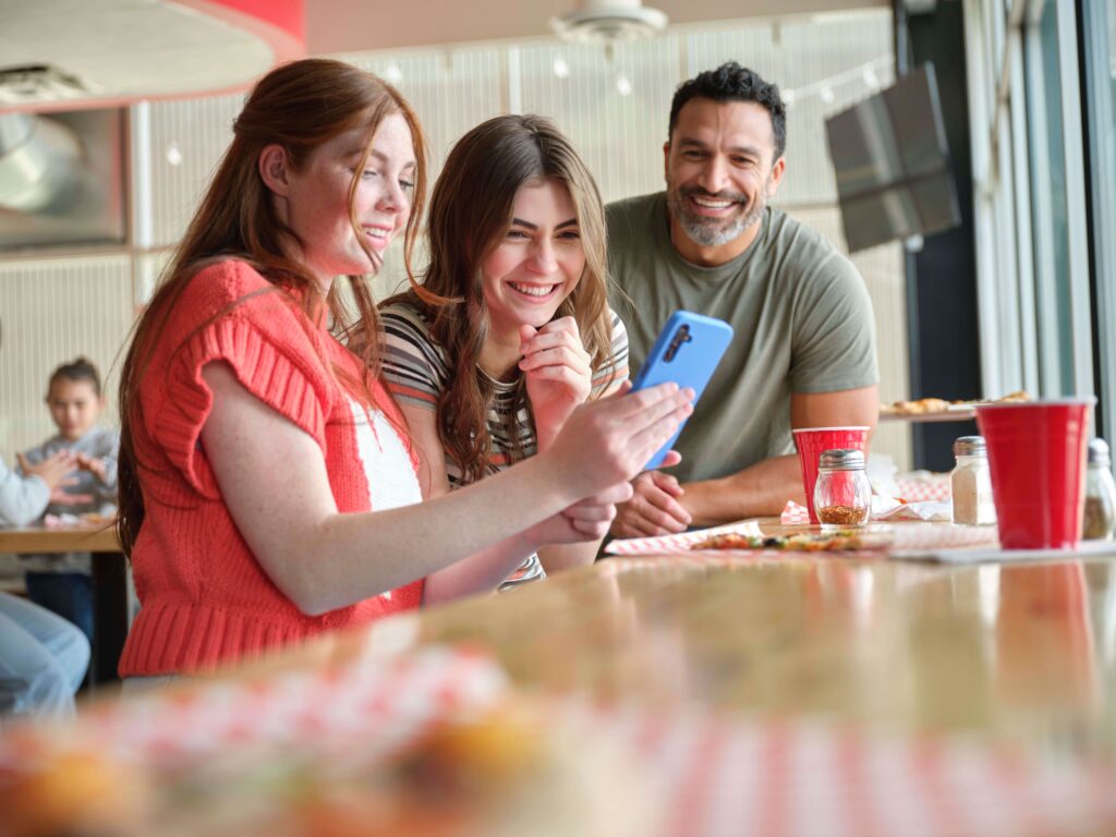 Two daughters with a phone with a dad observing at a restaurant smiling.