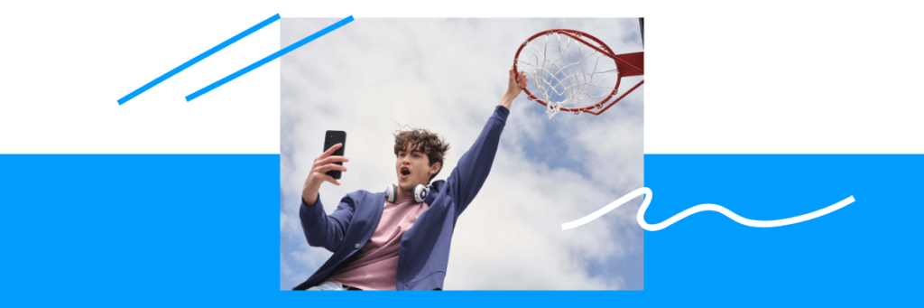 teen boy with cell phone hanging from a basketball hoop rim with blue cloudy sky behind him